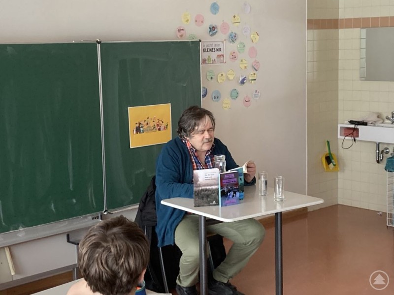 Ein &auml;lterer Mann sitzt an einem Lehrerpult vor einer Schultafel und liest aus einem Buch vor. Auf dem Tisch stehen zwei Gl&auml;ser und seine B&uuml;cher. Im Hintergrund sind Wanddeko und ein Waschbecken sichtbar.