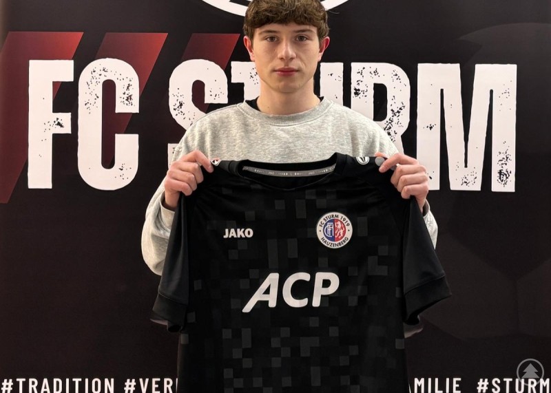 Jan Leimpek wechselt vom TSV Mauth zum FC Sturm Hauzenberg. Ein junger Fußballspieler hält ein schwarzes Trikot des FC Sturm Hauzenberg mit Sponsorenaufdruck vor einer Vereinswand mit Logo.
