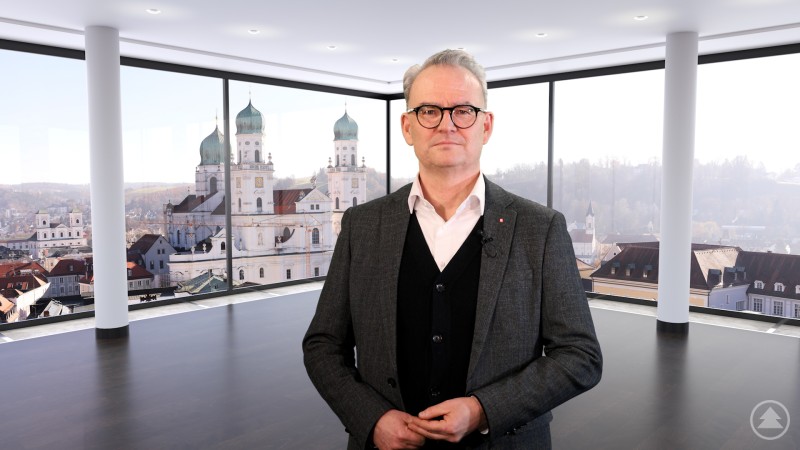Stefan Seiderer steht in einem hellen Raum mit Blick auf die Stadt Passau im Hintergrund und blickt in die Kamera.