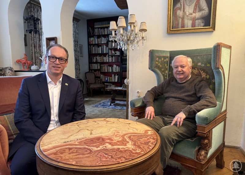 Das Schloss Buchenau empfängt mit einer großen Würde und Pracht. Allerdings ist es mit den Jahren auch zum Kostenfaktor für den Förderverein geworden. Dr. Roman Eder (r.) freute sich deshalb sehr, dass mit Mitteln der Freie Wähler Fraktionsreserve eine historische Erfassung möglich wird, um damit die nötigen Argumente für öffentliche Förderanträge sammeln zu können. Zwei Männer sitzen in einem historischen Innenraum an einem runden Tisch. Im Hintergrund sind Bücherregale, ein Kronleuchter und ein gerahmtes Gemälde zu sehen.