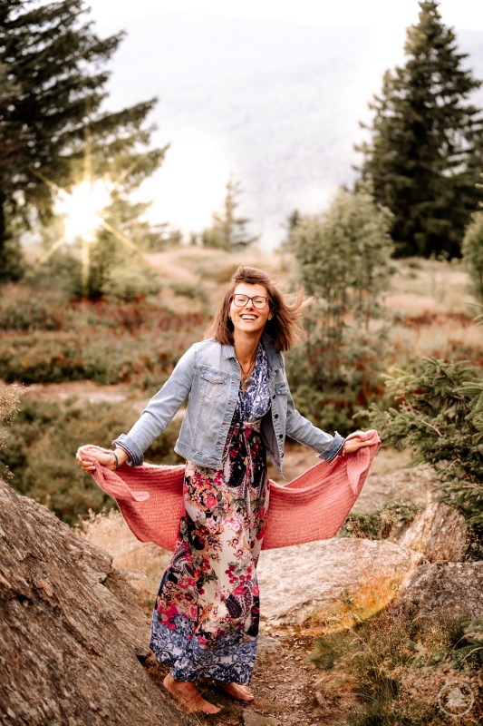 Andrea Wazlawiz hilft ihren Klienten ihren Seelenweg zu finden Andrea Wazlawiz mit braunem Schulterlangen Haar, Jeansjacke und Kleid vor einem natürlichen Hintergrund mit Bäumen und Gräsern. Sie lächelt strahlend und schwingt ihr Kleid.