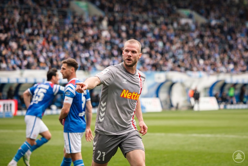 "Ein gelungener Tag" Ein Fußballspieler im grauen Trikot zeigt während des Spiels entschlossen mit dem Arm nach vorne, während im Hintergrund Gegenspieler in Blau und die Zuschauerränge zu sehen sind.