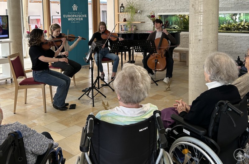 Musikerinnen und Musiker der European Orchestra Academy bringen klassische Musik direkt zu den Menschen, hier bei einem Konzert in einem Seniorenzentrum. Ein Streichquartett spielt in einem modernen Aufenthaltsraum eines Seniorenzentrums vor Zuhörern, die teilweise im Rollstuhl sitzen.