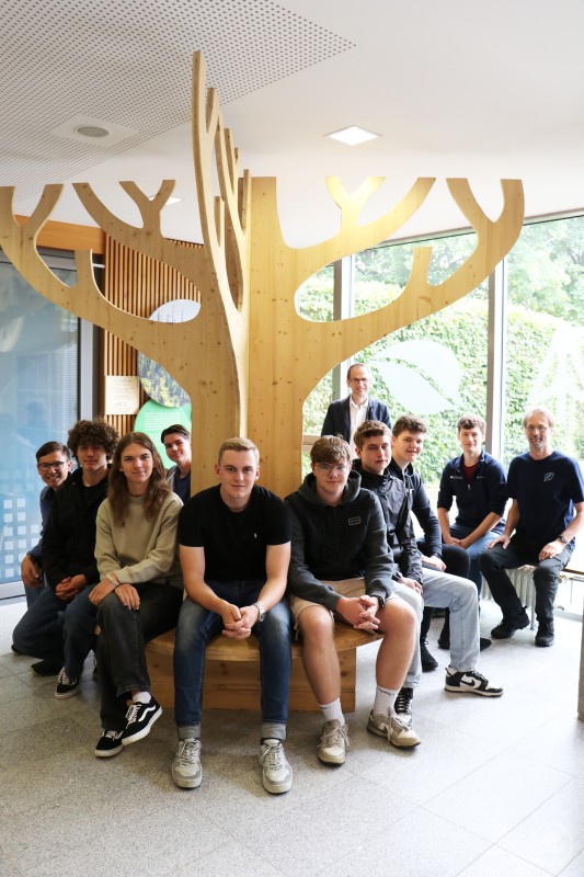 Eine Schülergruppe sitzt im Innenbereich der Schule auf einer Holzbank, die um eine große dekorative Holzbaumkonstruktion gebaut ist. Im Hintergrund stehen Lehrkräfte und Mitarbeitende des Nationalparks.