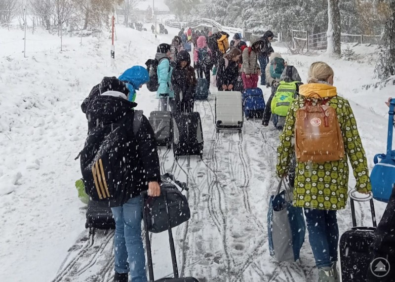 Kinder und Lehrkräfte ziehen ihre Koffer durch den Schneefall auf einem verschneiten Weg, um das Schullandheim zu erreichen. Dichte Schneeflocken und herbstliche Bäume prägen die Szenerie.