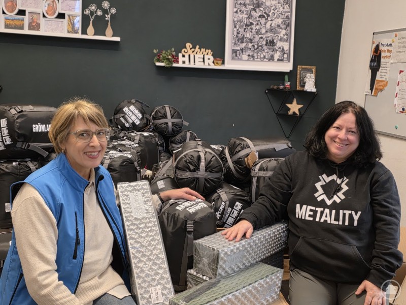 Renate Linner (links) vom Metality Chapter Passau übergibt 48 Schlafsäcke und 48 Isomatten an die Bahnhofsmission Passau. Zwei Frauen sitzen vor einem Stapel ausgerollter Isomatten und mehreren Schlafsäcken, die zur Spende bereitliegen. Beide lächeln in die Kamera, im Hintergrund sind Wanddekorationen und eine Tafel sichtbar. Die Frau rechts trägt einen schwarzen Hoodie mit „METALITY“-Logo, die Frau links eine blaue Weste.