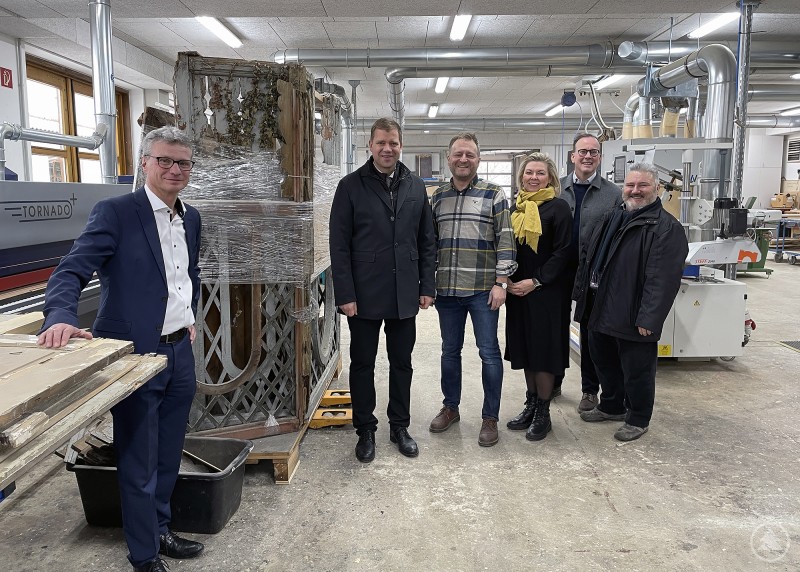 Landrat Bernd Sibler (v. l.), Bezirkstagspräsident Dr. Olaf Heinrich, Thomas und Bettina Hierbeck, Dr. Reinhard Saller, Geschäftsführer Niederbayern-Forum und Bürgermeister Alois Oswald. Sechs Personen stehen in einer modernen Schreinerei zwischen Maschinen und historischen Holzelementen, die auf Paletten gelagert sind. Im Hintergrund sind Maschinen und Werkstatteinrichtung sichtbar. Die Gruppe wirkt freundlich und professionell, alle blicken in die Kamera.