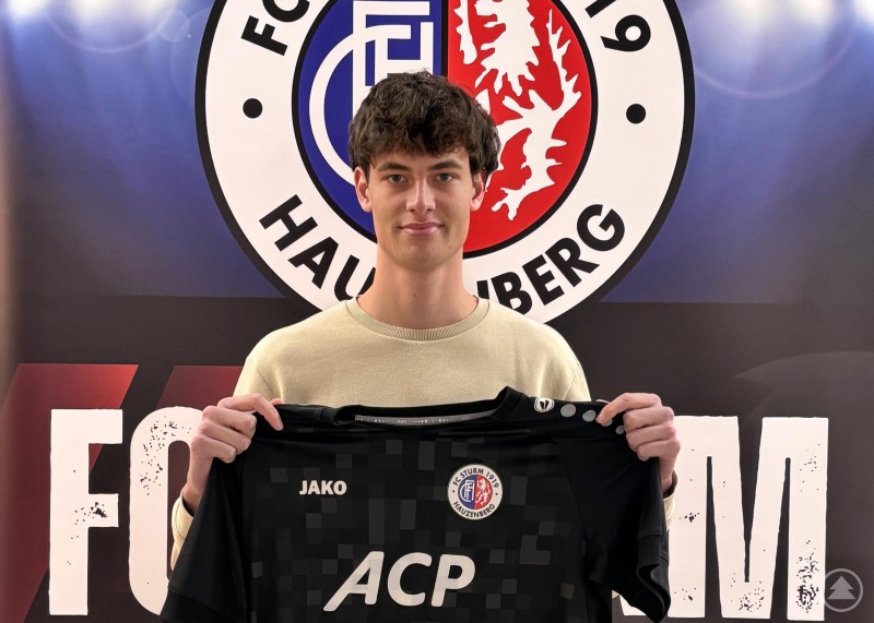 Julian Bauer kommt vom TSV Waldkirchen nach Hauzenberg. Ein junger Spieler steht mit einem Trikot des FC Sturm Hauzenberg vor einer Vereinswand und blickt in die Kamera.