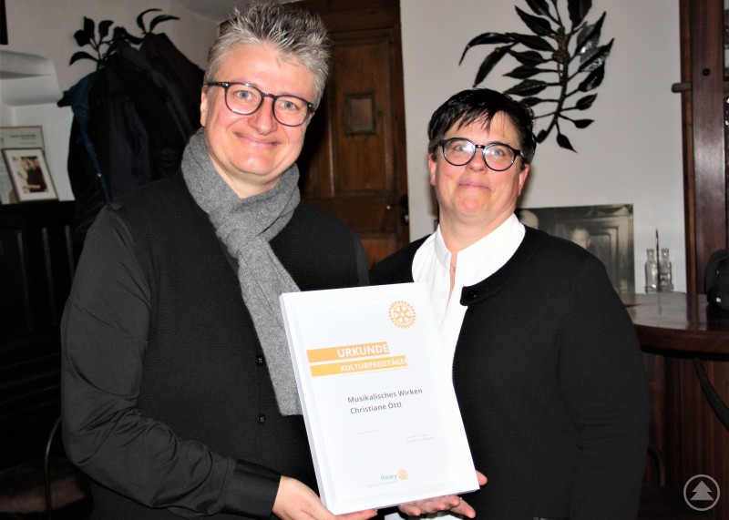 Die Urkunde zum mit 1000 Euro dotierten Musik-Kulturpreis des Rotary-Clubs Passau-Dreiflüssestadt konnte Christiane Öttl (links) in der Heilig Geist Stiftschänke von Club-Präsidentin Caroline Schober (rechts) entgegennehmen. Diese lobte das breit gefächerte musikalische und kulturelle Wirken der Preisträgerin. Zwei Frauen stehen nebeneinander in einem Gastraum und halten eine Urkunde in die Kamera. Eine der beiden trägt einen Schal, im Hintergrund sind Garderobe und Pflanzen zu sehen.