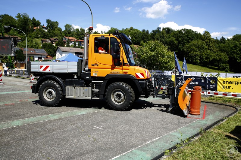 Ein Schneepflug schiebt drei aufrecht stehende, schwere Kunststoffrohre präzise in ein markiertes Rechteck auf Asphalt.