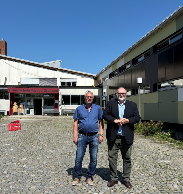 Oberbürgermeister Jürgen Dupper (rechts) begutachtet zusammen mit Anton Süß vom städtischen Hochbauamt die neue Fassade der Grundschule Passau-Grubweg. Zwei Männer stehen vor der sanierten Grundschule Passau-Grubweg; im Hintergrund sind die neue Fassade und das Schulgebäude zu sehen.