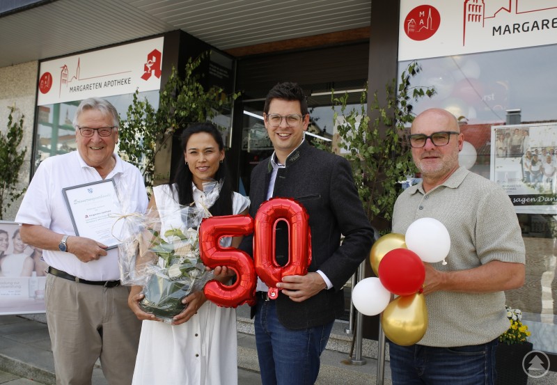 Peter Pleintinger gründete und führte die Margareten Apotheke bis zur Generationenübergabe an Tochter Elena. Zum 50. Geburtstag gratulierten Bürgermeister (2.v.r.) und Josef Lang (r.). Vier Personen stehen vor der Margareten Apotheke, darunter eine Frau mit Blumenstrauß und ein Mann mit Urkunde. In der Mitte werden rote Luftballons in Form der Zahl „50“ gehalten, rechts bunte Ballons. Im Hintergrund ist das Apothekenlogo sichtbar.