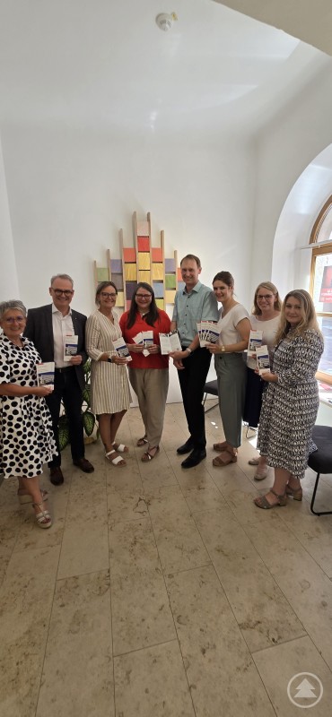 Caritas-Verantwortliche präsentieren die neue Spendenbroschüre für das Konradinum in Passau. Eine Gruppe von neun Personen steht in einem hellen Raum und hält Broschüren in den Händen. Im Hintergrund ist ein farbenfrohes Wandobjekt zu sehen.