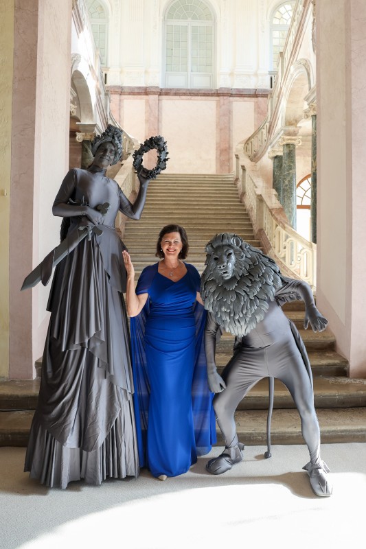 Auf Du und Du mit Löwe und Bavaria: Ilse Aigner lud zum Empfang und mehr als 3500 Gäste aus ganz Bayern schauten gerne vorbei. Eine Frau in königsblauem Kleid steht fröhlich zwischen zwei in Grau verkleideten Figuren – eine als stilisierte Bavaria mit Lorbeerkranz, die andere als Löwe mit Mähne – in einem historischen Treppenhaus.