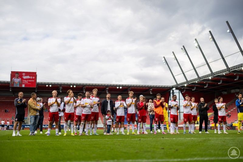 Die komplette Mannschaft steht nebeneinander auf dem Spielfeld, offenbar nach einem Spiel. Einige Spieler klatschen, andere haben Kinder oder Teammitglieder bei sich. Im Hintergrund sind die Tribünen des Stadions zu erkennen.