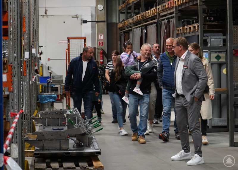 Gäste erhalten Einblicke in moderne Lager- und Produktionsprozesse. Eine Besuchergruppe geht durch ein Firmenlager mit hohen Regalen und Maschinen.