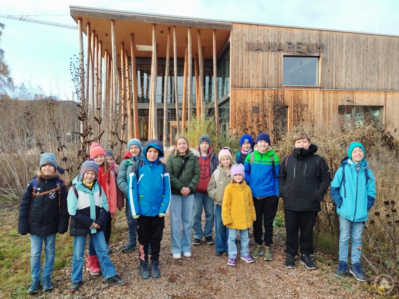 Gruppenfoto vorm Museum und in Aktion im Museum Eine Gruppe von Kindern und Jugendlichen steht draußen vor einem modernen Holzgebäude. Die Kinder tragen Winterkleidung und lächeln in die Kamera.