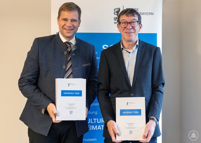Bezirkstagspräsident Dr. Olaf Heinrich präsentiert mit Bezirkskämmerer Winfried Amler (r.) den Haushalt 2026. Zwei Männer stehen nebeneinander vor einem Informationsbanner des Bezirks Niederbayern und halten jeweils ein gebundenes Dokument in den Händen. Beide lächeln in die Kamera. Es handelt sich um den Bezirkstagspräsidenten und den Bezirkskämmerer bei der Präsentation des Haushalts.