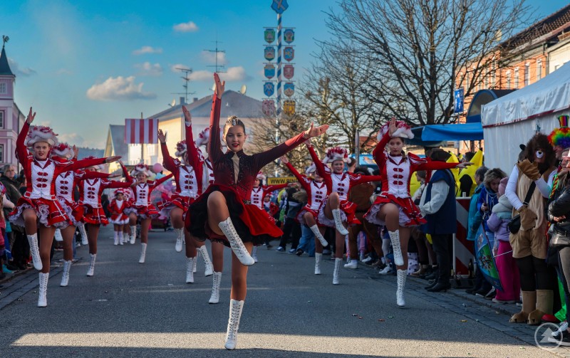 Garden und Faschingsgesellschaften sorgen beim Umzug am Marktplatz für Showtanz und Stimmung. Eine Garde in roten Uniformen marschiert und tanzt durch den Ort, im Vordergrund führt eine Tänzerin eine Pose aus, am Rand stehen Zuschauer.