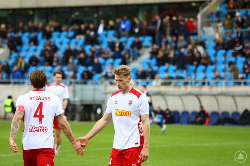 Zwei Spieler des SSV Jahn Regensburg klatschen sich w&auml;hrend eines Spiels auf dem Rasen ab, im Hintergrund sind Zuschauer auf der Trib&uuml;ne zu sehen.