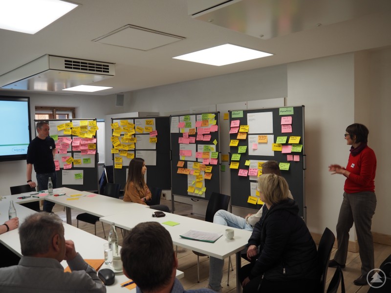 Beim Verstetigungsworkshop des difu zum Modellprojekt Smart City-SmartesLand im Ilzer Land brachten sich die Vertreter/-innen der teilnehmenden Gemeinden engagiert und kreativ ein. Das Bild zeigt eine kreative Workshop-Szene in einem modernen Besprechungsraum. An mehreren Pinnwänden hängen farbige Moderationskarten in Gelb, Pink, Grün und Orange – sie zeigen, dass hier intensiv Ideen gesammelt, geclustert und diskutiert werden. Eine Frau (rechts) spricht vor der Gruppe, während ein Mann (links) und mehrere Teilnehmende aufmerksam zuhören. Auf dem Tisch liegen ebenfalls Karten, Unterlagen und Wasserflaschen. Vermutlich handelt es sich um einen Strategie-Workshop oder eine Bürgerbeteiligung im Rahmen eines Projekts wie „Smart City“ oder kommunaler Entwicklung. Die Atmosphäre wirkt konstruktiv und offen für Austausch.