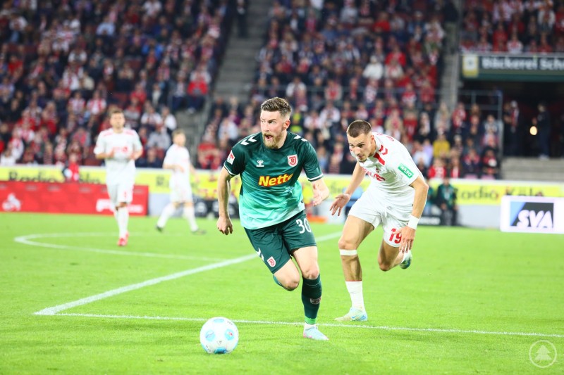 Spieler des SSV Jahn Regensburg im Zweikampf mit einem Kölner Angreifer – intensive Szenen im Abstiegskampf. Fußballspieler vom SSV Jahn Regensburg im Duell mit einem Spieler des 1. FC Köln während eines Spiels vor vollen Zuschauerrängen.