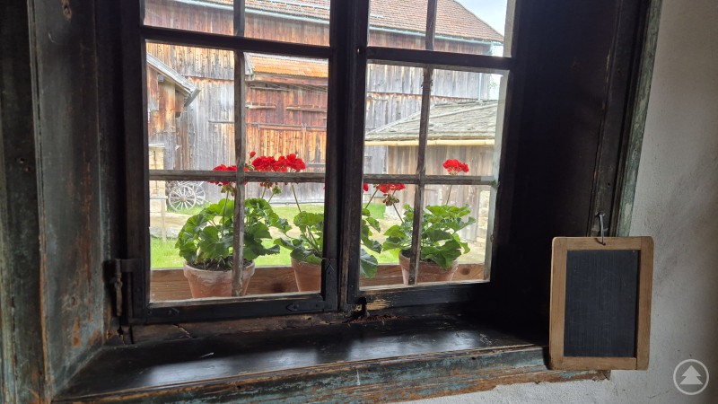 Blick aus dem Fenster vom Petzi Hof Blick durch ein altes Holzfenster mit Blumenkästen auf dem Sims. Draußen sind ein Bauernhof und ein rotes landwirtschaftliches Gerät zu sehen
