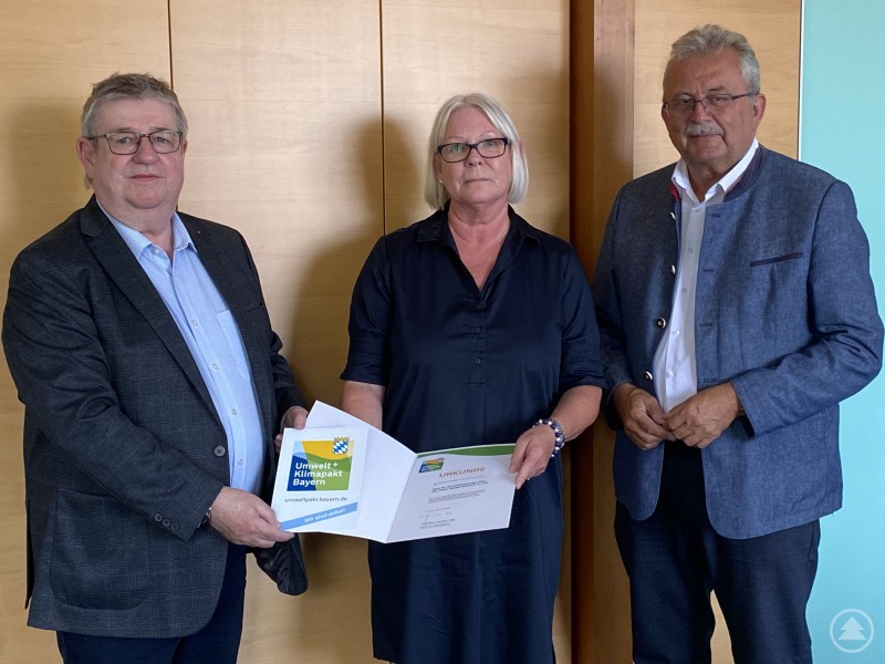 Dieter Hantke, Ulrike Schäfer und Landrat Josef Laumer (von links) bei der Übergabe der Urkunde im Büro des Landrats. Drei Personen stehen nebeneinander in einem Büro, zwei von ihnen halten eine Urkunde und eine Plakette mit dem Logo des Umwelt- und Klimapakts Bayern. Alle tragen Businesskleidung, im Hintergrund eine Holzvertäfelung.
