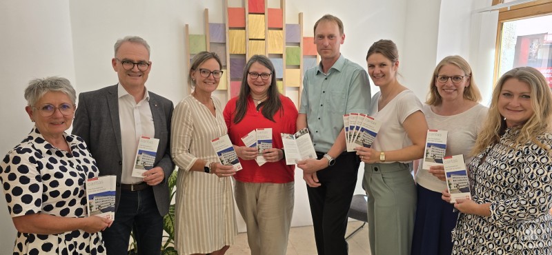 Gemeinsamer Einsatz für Menschen in Not: Caritas-Team mit der neuen Informationsbroschüre. Acht Personen stehen nebeneinander vor einer weißen Wand mit bunter Holzkunst. Sie halten Broschüren der Caritas in der Hand.