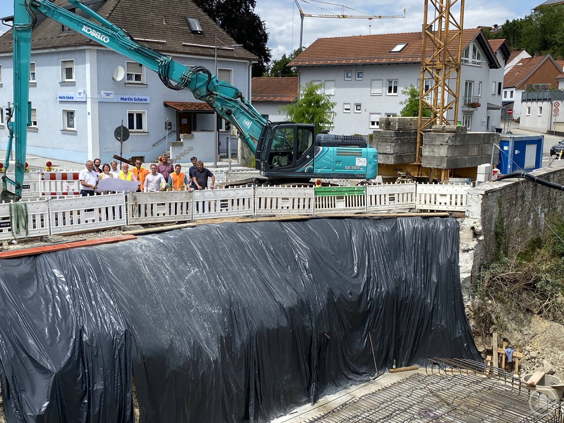 Ortstermin an der Baustelle der Bogenbachbrücke in Bogen – Politiker und Fachleute informieren sich über den aktuellen Baufortschritt. Eine Baustelle mit tiefem Aushub und schwarz abgedeckter Wand; darüber stehen Menschen an einem Bauzaun, im Hintergrund Bagger und Häuser.