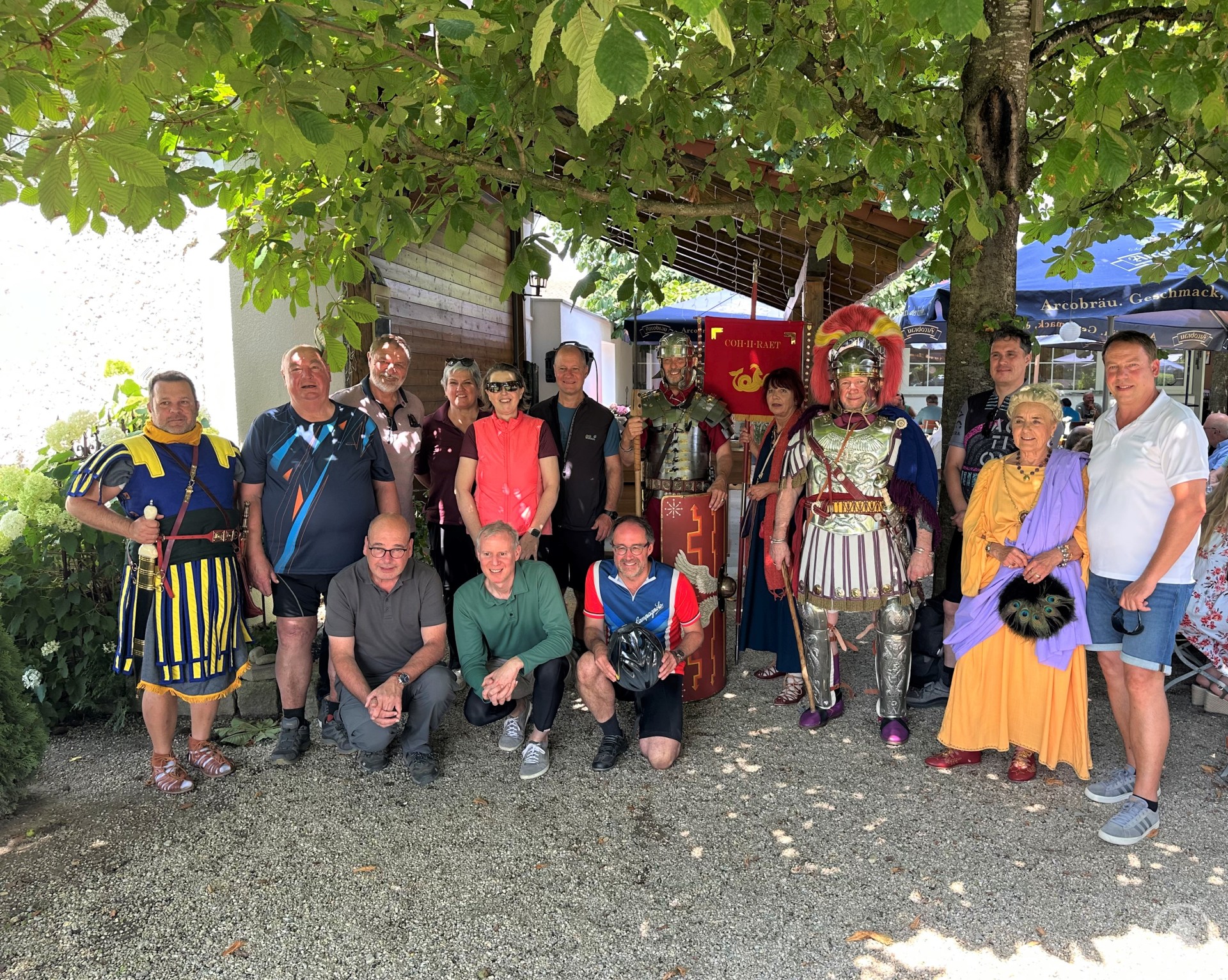 Eine Gruppe von rund 20 Personen, darunter Teilnehmer in historischer Römermontur sowie Alltagskleidung, steht unter einem Baum im Schatten. Im Hintergrund sind Schilder und ein römisches Banner zu erkennen.