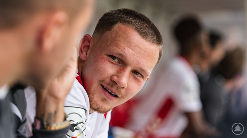 Ein junger Fußballspieler im Trikot des SSV Jahn Regensburg sitzt auf der Bank und lächelt im Gespräch mit einem Mitspieler.