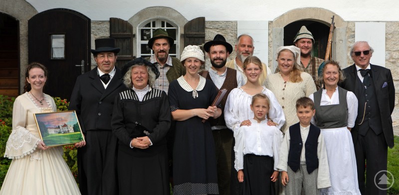 Eine größere Gruppe von Menschen posiert vor einem alten Gebäude mit weißen Mauern und Holzläden, alle tragen historische Kleidung, die an das 19. oder frühe 20. Jahrhundert erinnert. Zu sehen sind Frauen in langen Kleidern und Schürzen, Männer in Anzügen, Westen, Hüten und traditionellen Kopfbedeckungen sowie zwei Kinder in passenden Gewändern. Links hält eine Frau in einem hellen Kleid ein gerahmtes Bild in den Händen. Die Gruppe wirkt fröhlich und zusammengeschlossen, als handle es sich um eine nachgestellte historische Szene oder eine Brauchtumsveranstaltung.