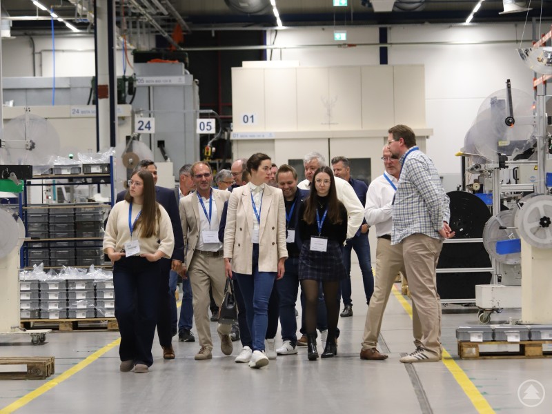 Mitten in der Produktionshalle des Gastgebers konnten sich die Teilnehmerinnen und Teilnehmer nicht nur ein Bild der Prozesse bei Hirschmann Automotive machen, sondern sich auch über konkrete Anwendungsfelder Künstlicher Intelligenz informieren. Eine Gruppe von Teilnehmerinnen und Teilnehmern geht durch eine Produktionshalle bei Hirschmann Automotive. Sie tragen Namensschilder und hören einem Mitarbeiter zu, der ihnen Prozesse und Abläufe erklärt. Im Hintergrund sind moderne Maschinen und Produktionsanlagen zu sehen.