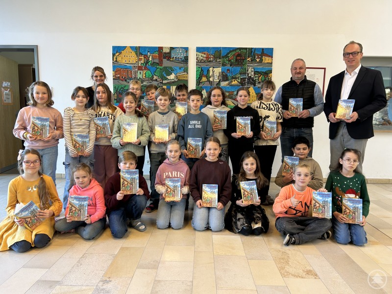 Die Schülerinnen und Schüler der Reinhold-Koeppel-Grundschule Grafenau mussten sich nicht extra um einen Klassensatz der Isar-Detektive bewerben. 35 Exemplare wurden nun über MdL Martin Behringer an Schulleiter Christoph Stockinger und Klassleiterin Eva-Maria Salzinger der 4a übergeben. Sie dürfen gierig verschlungen und der Landtag indirekt kennengelernt werden. Eine Schulklasse mit Kindern und zwei Erwachsenen steht und sitzt in einem Flur vor einer Wand mit bunten Bildern. Viele halten jeweils ein Buch in der Hand.