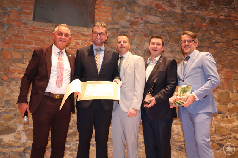 F&uuml;nf M&auml;nner in Anz&uuml;gen posieren vor einer Steinwand, einer h&auml;lt eine Urkunde, ein anderer eine Weinflasche, ein weiterer ein Buch mit dem Titel 9 Pl&auml;tze 9 Sch&auml;tze.