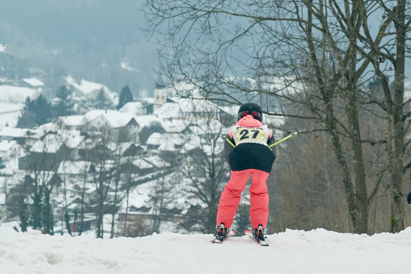 Ein Skifahrer mit Startnummer 27 f&auml;hrt eine verschneite Abfahrt hinunter, im Hintergrund sind H&auml;user im Tal zu sehen.