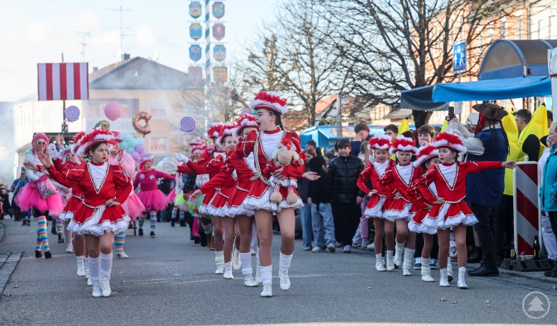 Beim Gaudiwurm ziehen die angemeldeten Gruppen mit Musik, Konfetti und kreativen Kostümen durch den Ortskern. Mehrere Gardetänzerinnen in roten Kostümen laufen in Formation durch die Straße, im Hintergrund sind weitere Gruppen und Feststimmung zu sehen.