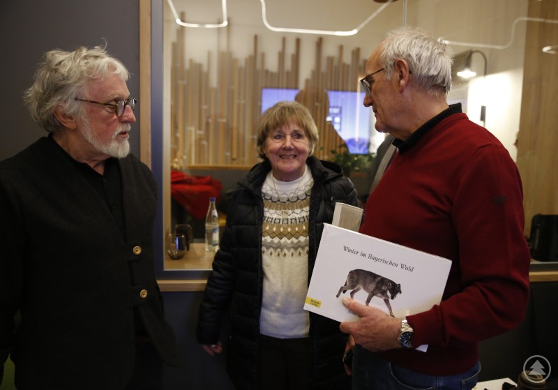 Heinz Glashauser mit Elisabeth Kern und stellvertretendem Bürgermeister Günter Klampfl unterhielten sich nach der Buchvorstellung über alte Zeiten im Markt und neue Bücher aus dem Bayerischen Wald. Drei Personen stehen in einem modern gestalteten Raum beisammen und unterhalten sich. Einer der Männer hält ein Buch mit dem Titel Winter im Bayerischen Wald in der Hand, auf dem ein Wolf abgebildet ist.