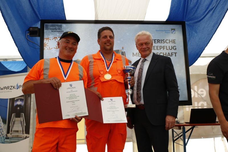 Die Straßenmeisterei Vilshofen holt den Titel: Bayerischer Schneepflugmeister 2025 in Schönberg. Ein orangefarbener Unimog der Straßenmeisterei Vilshofen steht mit Schneepflug vor Publikum auf dem Sportgelände.