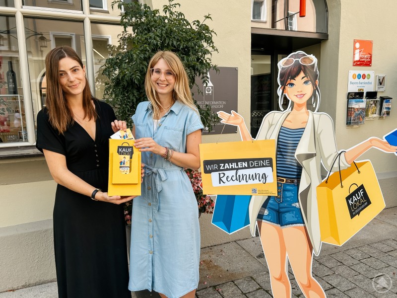 Die Rechnungen können in die Einwurf-Box in der Tourist Information im Alten Rathaus eingeworfen werden. Foto: Stadtmarketing Deggendorf e.V. Zwei junge Frauen stehen vor einem Gebäude mit Schaufenstern und einem Eingangsschild „Tourist Information“. Sie halten eine gelbe Box mit dem Aufdruck „Kauf Lokal“. Daneben steht ein lebensgroßer Pappaufsteller einer gezeichneten Frau mit Einkaufstaschen und dem Hinweis: „Wir zahlen deine Rechnung“.