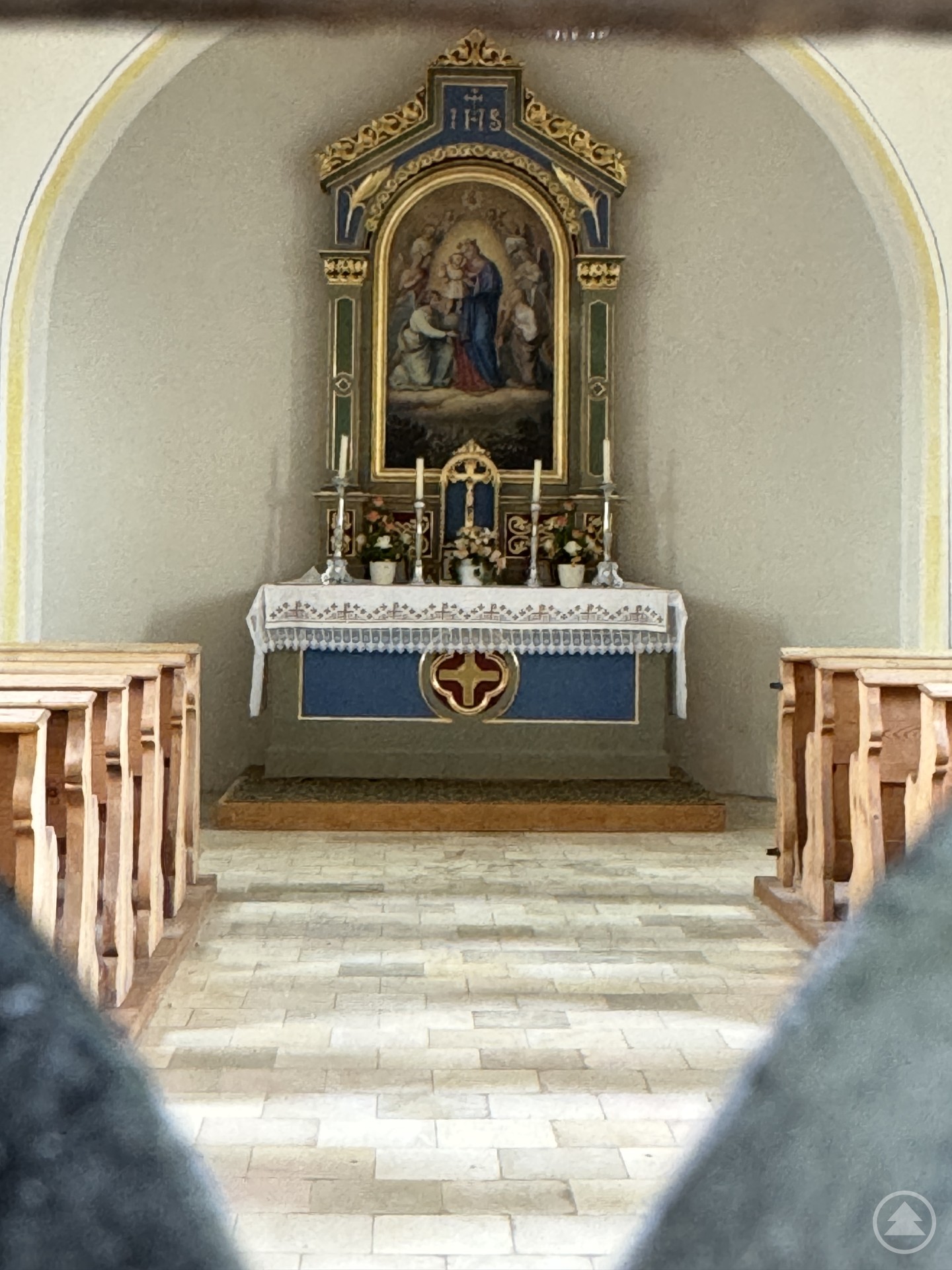 Blick durch zwei Bankreihen auf den Altarraum einer kleinen Dorfkapelle mit klassizistischem Altarbild und Blumenschmuck.