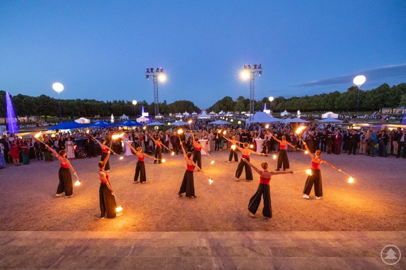 Eine Gruppe von Künstlerinnen in rot-schwarzen Kostümen führt bei Einbruch der Dunkelheit eine Feuershow im Schlosspark vor großem Publikum auf. Im Hintergrund leuchten blaue Fontänen und Lichterzelte.