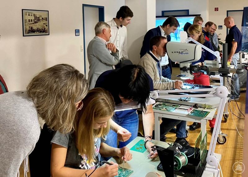 Kinder und Erwachsene löten und tüfteln gemeinsam an elektronischen Bauteilen. Mehrere Personen, darunter Kinder, sitzen an Arbeitstischen und probieren sich an Löt- und Elektronikarbeiten.