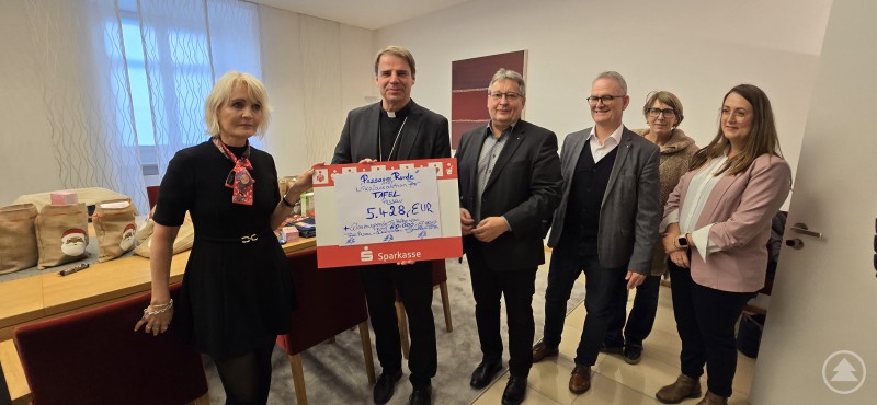 Eine herausragende Hilfe für Menschen in Not. Claudia Gugger-Bessinger, überreicht Bischof Dr. Stefan Oster den Scheck über 5428 Euro. Mit dabei Diakon Konrad Niederländer, sowie dessen Nachfolger als Caritasvorstand Stefan Seiderer, Angelika Leitl-Weber (Bahnhofsmission) und Monika Winkler (Tafel Passau). Mehrere Personen stehen in einem Besprechungsraum zusammen und präsentieren einen großen Spendenscheck.