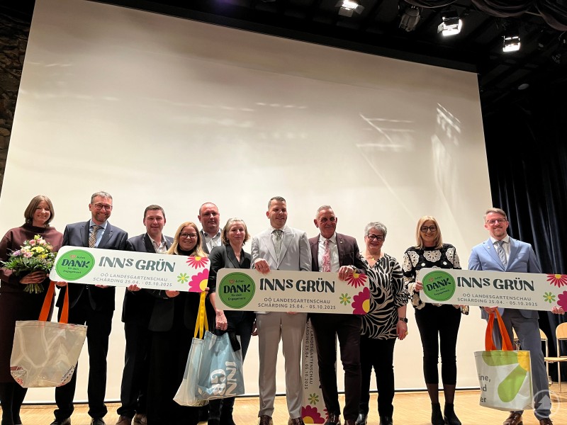 Gruppe von Frauen und M&auml;nnern auf einer B&uuml;hne, sie halten gro&szlig;e Schilder mit INNS GR&Uuml;N und tragen Geschenktaschen, im Hintergrund eine gro&szlig;e Leinwand.