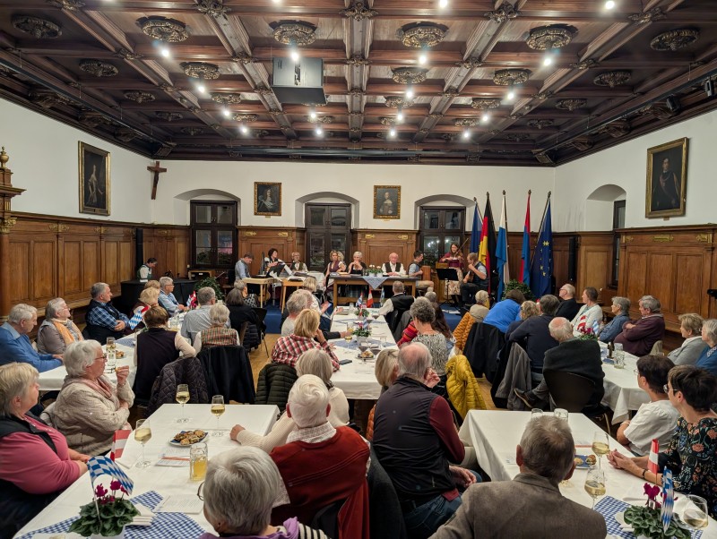 Der Dreierlei Mundart Hoagarten im Alten Rathaus war in den letzten beiden Jahren bis auf den letzten Platz gefüllt und sorgte für großen Applaus. Im Historischen Saal des Alten Rathauses sitzen zahlreiche Besucherinnen und Besucher an festlich gedeckten Tischen. Vorne auf einer kleinen Bühne musizieren und lesen mehrere Künstler, während bayerische und europäische Fahnen im Hintergrund stehen.