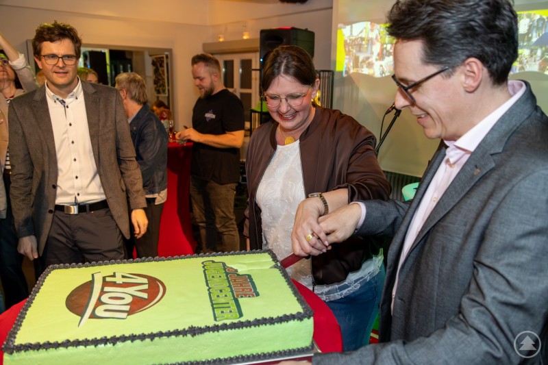 Oberb&uuml;rgermeister Dr. Christian Moser und Jugendcenter-Leiterin Karin Loibl schneiden gemeinsam eine gro&szlig;e Geburtstagstorte zum 18-j&auml;hrigen Bestehen des Jugendcenters 4You an, w&auml;hrend G&auml;ste die Feier beobachten.