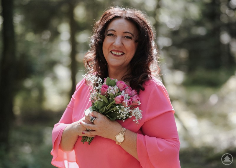 Jeanette Maier hat langes schwarzes Haar, tr&auml;gt ein pinkes Shirt und h&auml;lt einen fr&ouml;hlichen pinken Blumenstrau&szlig; vor Waldhintergrund in die Kamera.