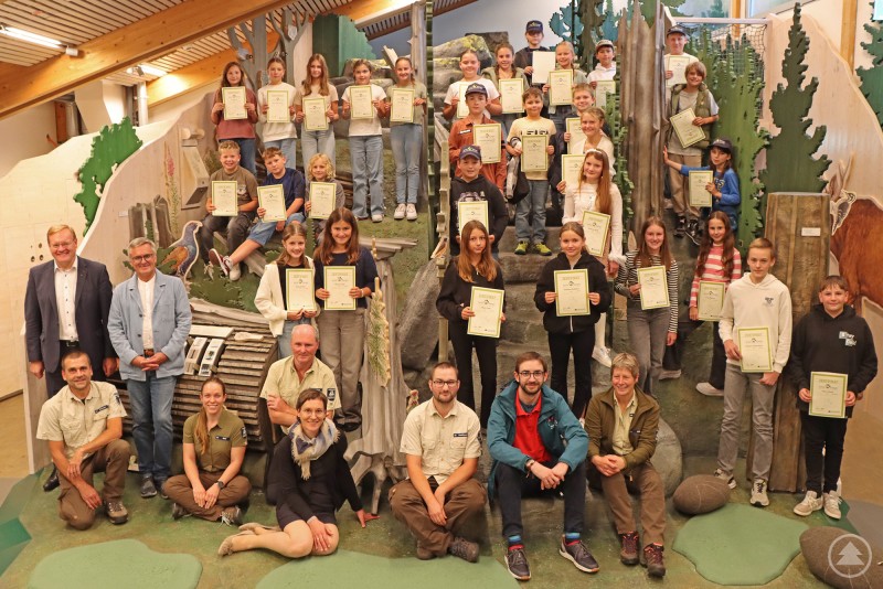 Zum wilden Gruppenbild stellten sich nach der kurzen Feierstunde die neuen Junior Ranger nebst Ehrengästen in der Waldwerkstatt auf. Eine große Gruppe von Kindern und Jugendlichen posiert mit Urkunden in den Händen gemeinsam mit Betreuern und Ehrengästen in einem Raum der Waldwerkstatt des Nationalparks Bayerischer Wald.